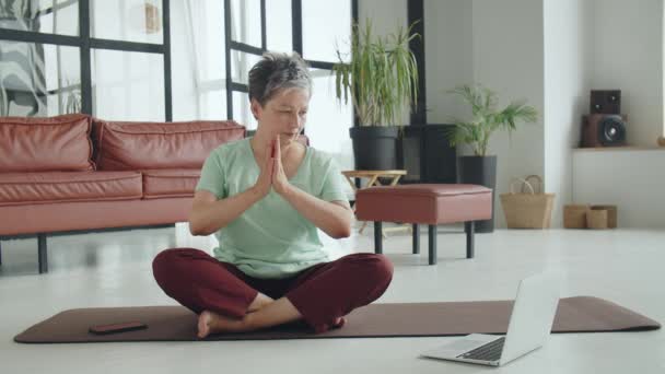 Old woman make yoga exercise at home. Mature woman have meditation. She look at video on laptop online courses. High quality 4k footage - Footage, Video