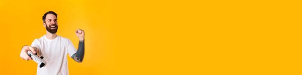 Felice uomo barbuto bruna di mezza età che tiene il joystick in piedi sopra il muro giallo, celebrando il successo - Foto, immagini