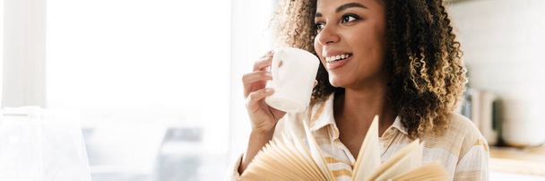 La femme noire heureuse avec tasse et livre dans ses mains regardant sur le côté tout en étant assis à la table dans la cuisine légère - Photo, image