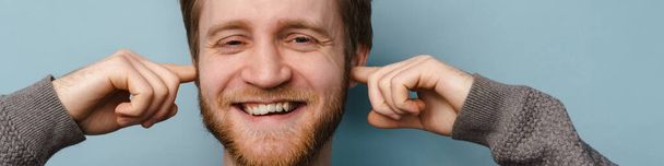 Bearded ginger man smiling while plugging his ears isolated over blue background - Фото, изображение