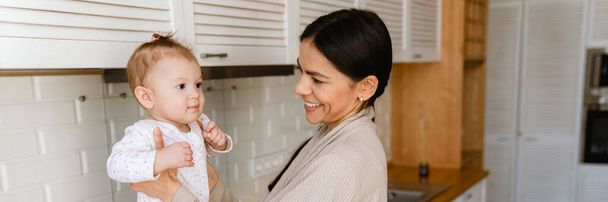 Joven madre sonriendo y jugando con su bebé en la cocina en casa - Foto, Imagen