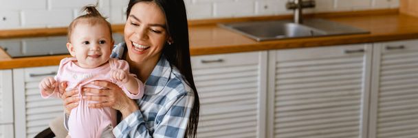Young mother smiling and holding her baby while working with laptop in kitchen at home - Фото, зображення