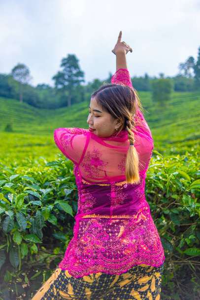 an Asian woman in a pink costume is posing in front of a tea plantation in the morning - Fotó, kép
