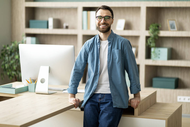 Porträt eines hübschen jungen Geschäftsmannes, der neben dem Schreibtisch im Büroinnenraum steht, selbstbewusster männlicher Millennial-Unternehmer in lässiger Kleidung, der am Arbeitsplatz posiert, in die Kamera blickt und lächelt, Kopierraum - Foto, Bild
