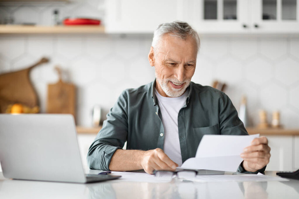 Счастливый пожилой человек, читая письмо, сидя за рабочим столом с верхушкой в Китчене, получил хорошие новости по почте от страховой компании, получил пенсию по выслуге лет - Фото, изображение