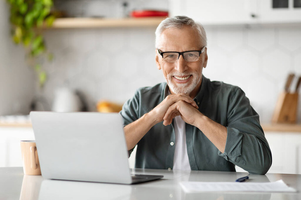 Freelance vanhemmalla iällä. Happy Senior Man istuu työpöydällä kannettavan tietokoneen kanssa keittiön sisustuksessa, komea vanhusten herrasmies käyttää tietokonetta online-työhön kotoa, hymyilee ja katselee kameraa - Valokuva, kuva
