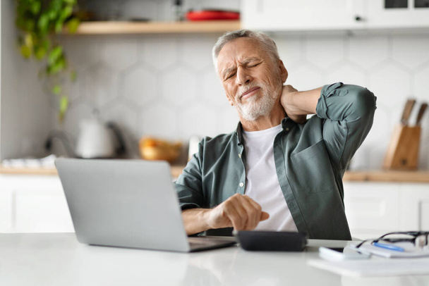 Κουρασμένος ηλικιωμένος άνθρωπος που υποφέρει από πόνο στο λαιμό μετά την εργασία με το laptop στο κεντρικό γραφείο, αναστατωμένος ανώτερος κύριος μασάζ φλεγμονή περιοχή και συνοφρύωμα, αίσθημα αδιαθεσία, ενώ κάθεται στο γραφείο στην κουζίνα - Φωτογραφία, εικόνα