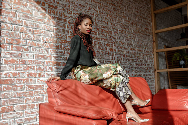 Όμορφο κορίτσι από την Αφρική που φοράει κομψά ρούχα, κάθεται στο πίσω μέρος του κόκκινου δερμάτινου καναπέ στο δωμάτιο σε έναν τοίχο από τούβλα. Ο φωτισμός της ηλιακής δέσμης πέφτει σε πρόσωπο, καναπέ και τοίχο. Αντιγραφή χώρου για κείμενο. - Φωτογραφία, εικόνα