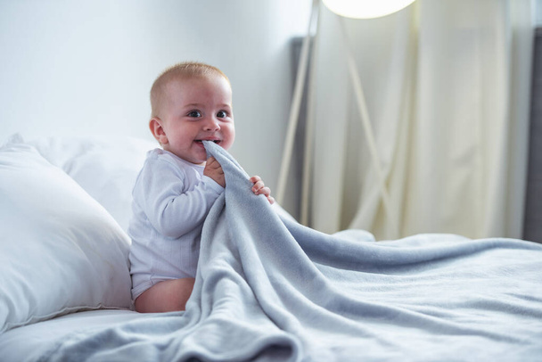 Charming happy baby is chewing a blanket, looking at camera and smiling while sitting on bed at home - Photo, Image