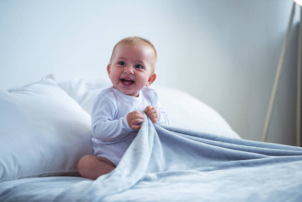 glücklich bezauberndes Baby hält eine Decke in der Hand, schaut weg und lacht, während es zu Hause auf dem Bett sitzt - Foto, Bild