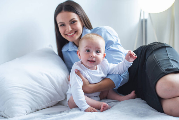 Бизнес-леди в классическом костюме держит своего милого малыша, смотрит в камеру и улыбается, лежа дома на кровати
 - Фото, изображение