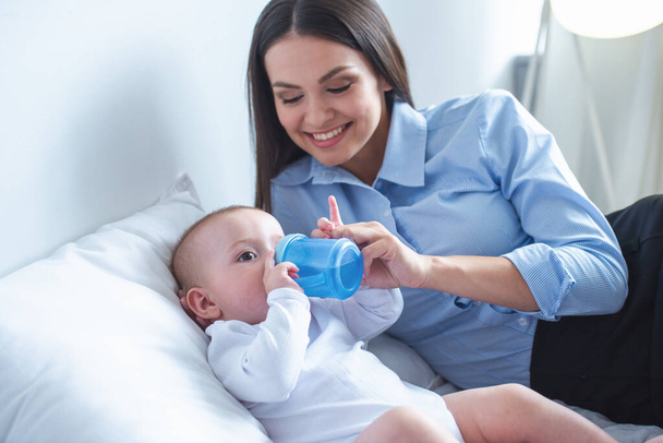 Charming little baby is drinking while lying near his business mom on bed at home - Photo, Image
