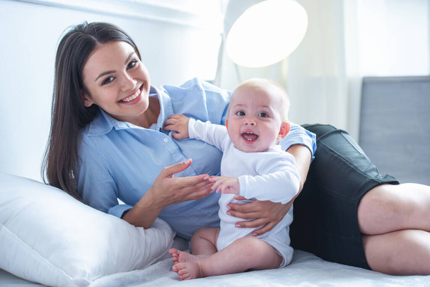 Belle femme d'affaires en costume classique et son doux petit bébé regardent la caméra et sourient tout en passant du temps ensemble sur le lit à la maison
 - Photo, image