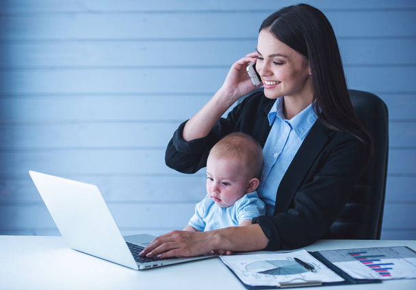 Bela senhora de negócios em terno clássico está falando no telefone celular e sorrindo, trabalhando no escritório enquanto seu bebê bonito está sentado de joelhos
 - Foto, Imagem