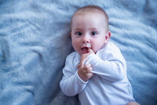 Muotokuva viehättävästä vauvasta, joka pitää sormea suussaan, katsoo kameraan ja hymyilee, makaa sängyssä kotona
 - Valokuva, kuva