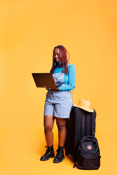 Female tourist using laptop in studio, waiting to leave on holiday trip. Young woman with trolley bags and luggage holding pc, travelling abroad and sitting over orange background. - Photo, Image