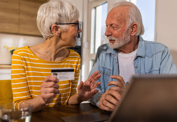 Joyeux couple de personnes âgées assis à table à manger avec cuisine en arrière-plan et achat en ligne avec carte de crédit  - Photo, image