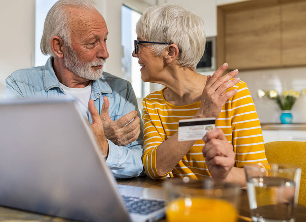 陽気な高齢者のカップルは、バックグラウンドでキッチンとダイニングテーブルに座って、クレジットカードでオンラインで購入  - 写真・画像
