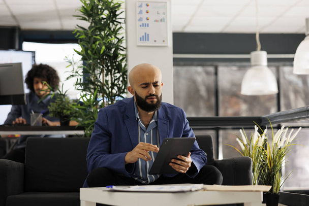 Arabische executive manager controleren van de financiële gegevens op tablet tijdens het werken in het kantoor. Startup bedrijf werknemer surfen op internet op digitaal apparaat en zitten op de bank in coworking ruimte - Foto, afbeelding