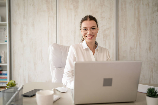 Beyaz gömlekli, beyaz gömlekli, iş kadını ya da sekreterli bir kadın bilgisayarının masasında oturuyor, gülümsüyor. - Fotoğraf, Görsel