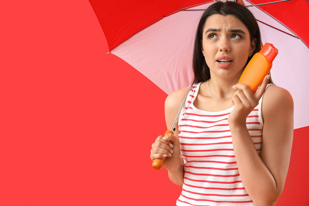 Worried young woman with sunscreen cream and beach umbrella on red background - Photo, Image