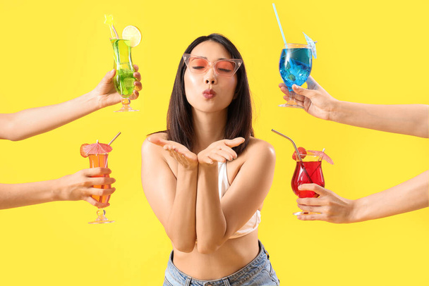 Beautiful Asian woman blowing kiss and hands with many cocktails on yellow background - Photo, Image