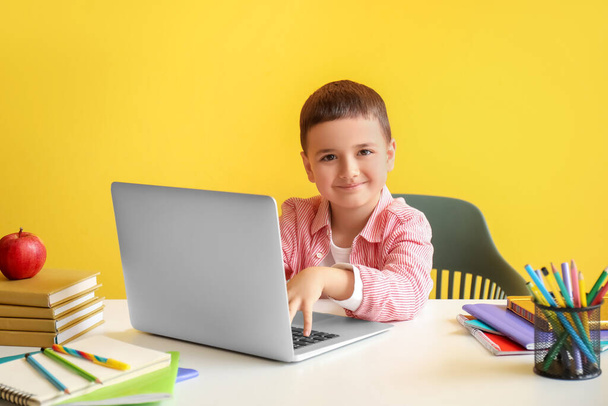 Χαριτωμένο μικρό αγόρι με φορητό υπολογιστή κάνει την εργασία στο τραπέζι κοντά στον κίτρινο τοίχο - Φωτογραφία, εικόνα
