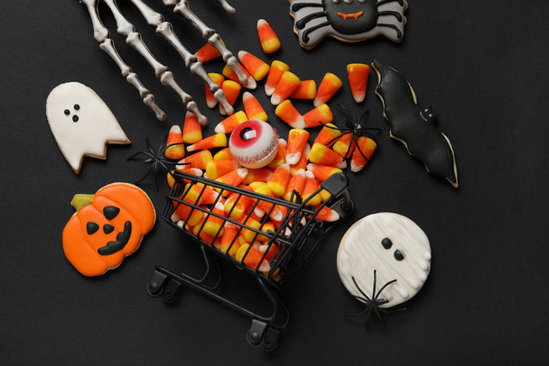 Carrito de compras con sabrosos callos de caramelo, esqueleto de mano y galletas para Halloween sobre fondo negro - Foto, imagen