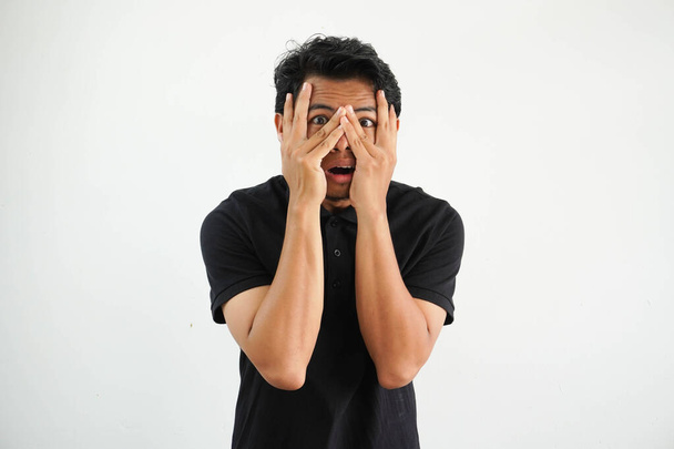 Молодий азіатський чоловік ізольований на білому тлі блимає крізь пальці переляканий і нервовий, одягнений в чорну футболку поло. - Фото, зображення