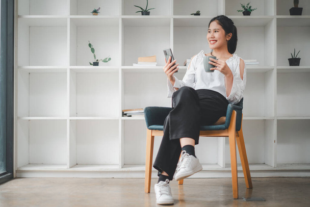 Szczęśliwa ładna azjatycka kobieta relaksująca się w domu, kobieta korzystająca z telefonu komórkowego, czatująca w sieci, pracująca lub studiująca online, siedząca w fotelu z przestrzenią do kopiowania. - Zdjęcie, obraz