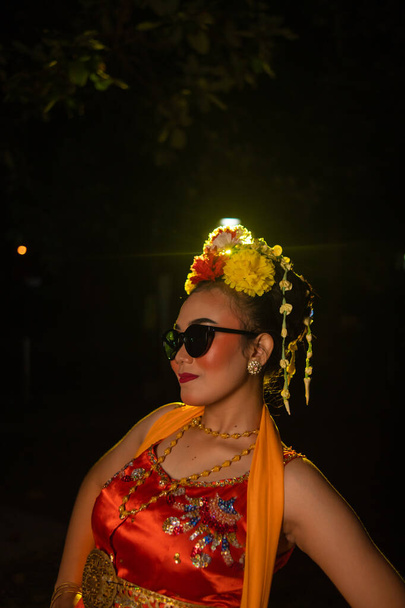 una ballerina sundanese posa davanti a una luce splendente indossando un costume arancione con occhiali da sole e fiori in testa di notte - Foto, immagini