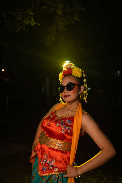 una ballerina sundanese posa davanti a una luce splendente indossando un costume arancione con occhiali da sole e fiori in testa di notte - Foto, immagini