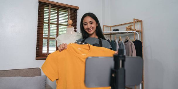Nuori aasialainen nainen yrittäjä tarkistaa tuotteen ja puhuva kamera elää tallennus video sosiaalinen verkosto kotona. Verkkomyynti, liiketoiminta ja teknologia. - Valokuva, kuva