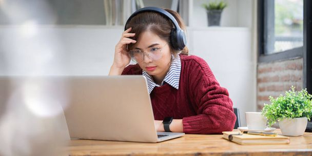 Asian college student at home wearing red shirt using laptop attending online university class listening with headphones. - 写真・画像