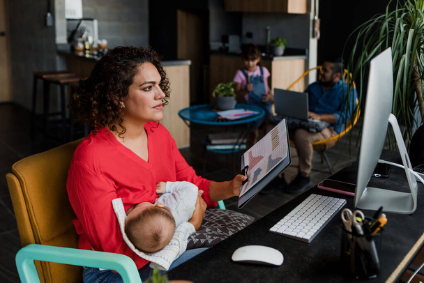 молодая латинская мать работает дома с помощью компьютера, пока она кормит грудью своего маленького сына в Мексике Латинская Америка, домашний офис концепции, испаноязычная семья - Фото, изображение