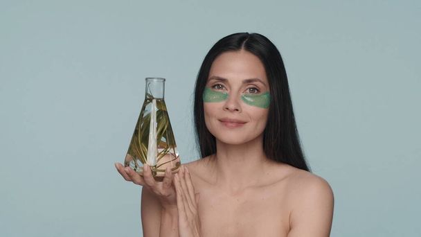 A woman with green hydrogel patches under her eyes holds a flask with a plant. Seminude woman in the studio on a blue background. Cosmetic products based on natural plant ingredients. The concept of - Valokuva, kuva