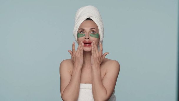 A woman wrapped in bath towels after a shower, with patches under her eyes and on her lips in a studio on a blue background. The woman smoothes the red patches on her lips. The concept of beauty - Foto, Imagem