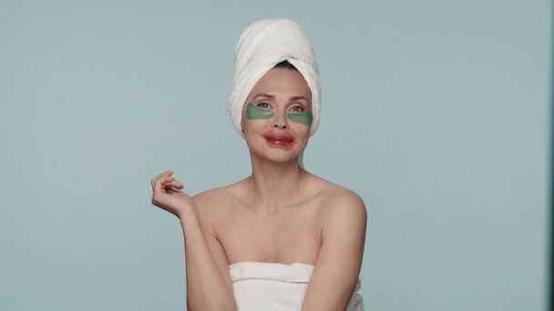Portrait of a woman wrapped in bath towels in the studio on a blue background close up. A woman performs cosmetic procedures after a shower, applies hydrogel patches for eyes and lips. - Photo, image