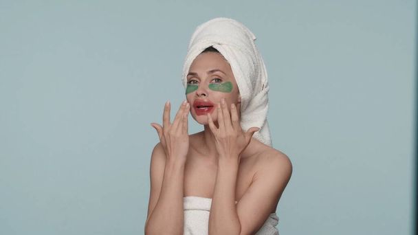 A woman wrapped in bath towels after a shower, with patches under her eyes and on her lips in a studio on a blue background. The woman smoothes the red patches on her lips. The concept of beauty - Valokuva, kuva