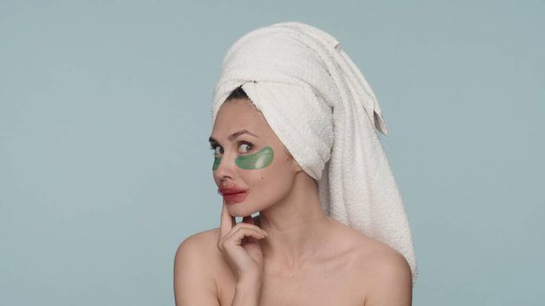 Portrait of a seminude woman with a towel on her head in the studio on a blue background. A woman performs cosmetic procedures after a shower, applies hydrogel patches on her eyes and lips. The - Fotografie, Obrázek