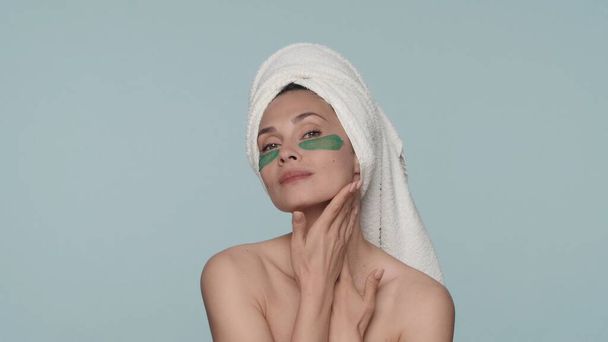 Portrait of a woman with a towel on her head and green hydrogel patches under her eyes close up. A woman enjoys the purity and smoothness of her skin in the studio on a blue background. The concept of - Foto, Imagem