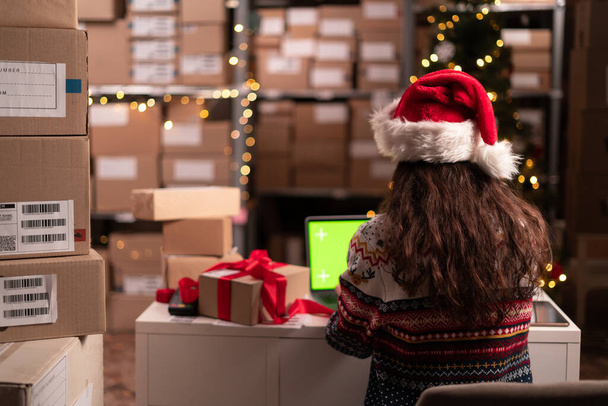 Managerin mit Weihnachtsmütze mit grünem Laptop-Attrappen-Computer, um Inventar zu überprüfen. Warehouse Einzelhandelszentrum mit Boxen im Weihnachtsverkauf, e-Commerce Online-Bestellungen. Kopierraum - Foto, Bild