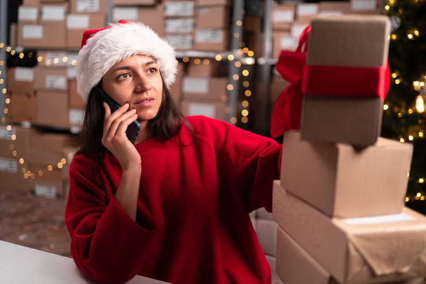 Wholesale, logistic, business, export and people concept. Female inventory manager with cardboard boxes calling on smartphone at warehouse. Christmas sale time - Photo, Image