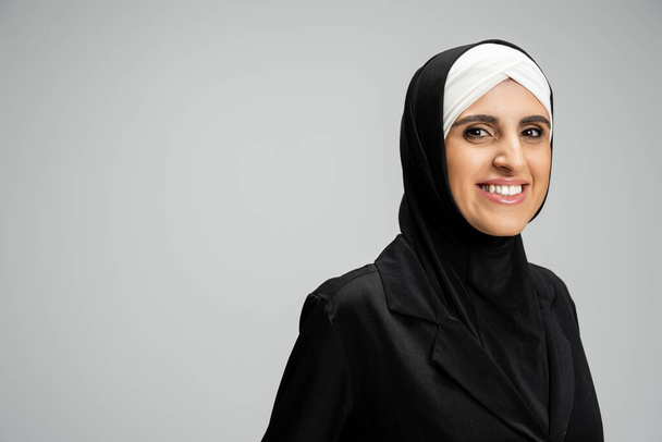 feliz musulmana mujer de negocios en hijab y chaqueta negra sonriendo a la cámara en gris, disparo en la cabeza - Foto, imagen