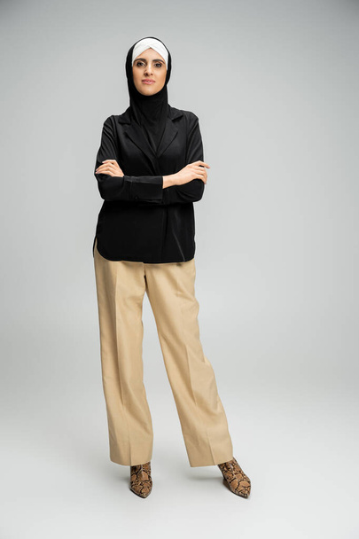 mujer de negocios musulmana con estilo en blazer, pantalones y hijab posando con los brazos cruzados en gris, longitud completa - Foto, Imagen