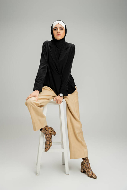 businesswoman in muslim hijab and trendy casual attire sitting on high stool on grey, full length - Foto, Imagen