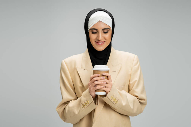 pleased middle eastern woman in muslim hijab and beige blazer holding coffee to go on grey - Foto, immagini