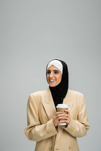 mujer musulmana positiva en chaqueta elegante y hijab sosteniendo taza de papel y mirando hacia otro lado en gris - Foto, imagen