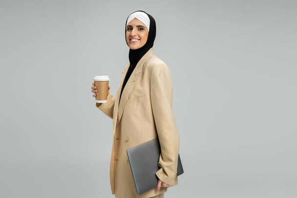 femme d'affaires musulmane moderne et élégante avec café à emporter et ordinateur portable souriant à la caméra sur gris - Photo, image