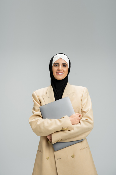 portrait of smiling muslim businesswoman in blazer and hijab holding laptop on grey, headshot - Photo, Image
